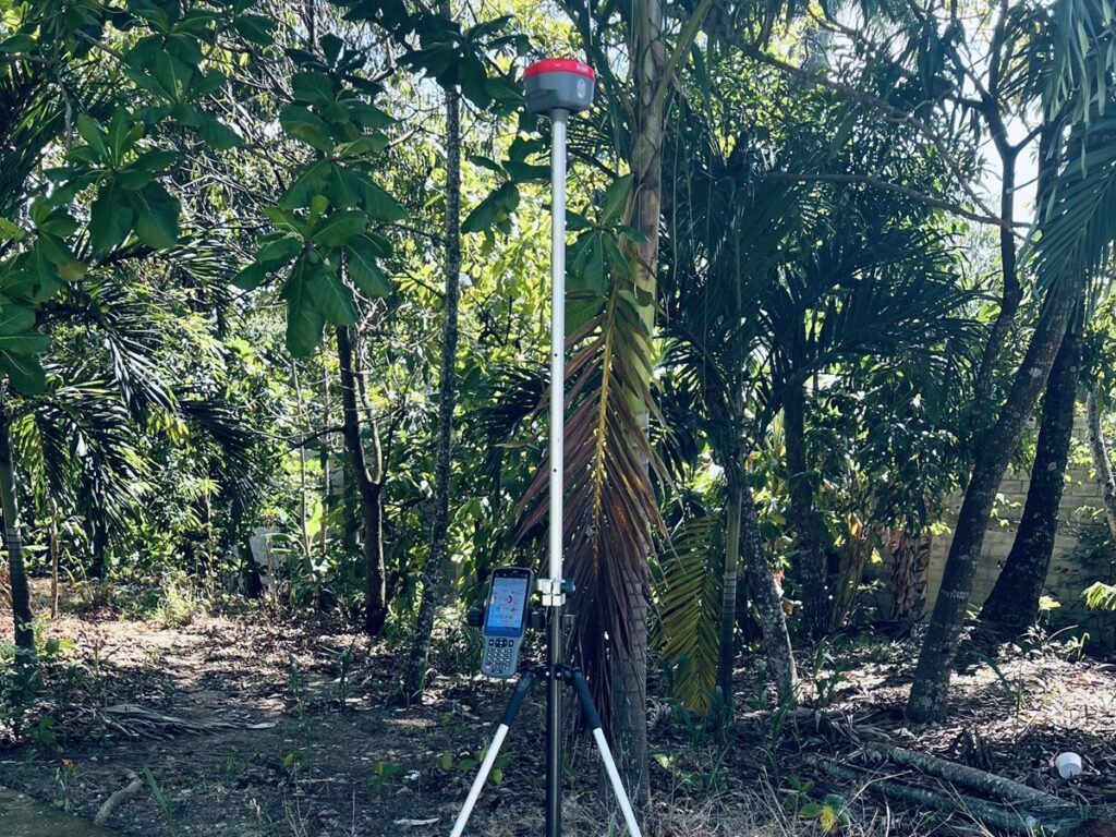 Receptor GNSS sobre bastón con apoyo y colectora, junto a un bordillo y vegetación