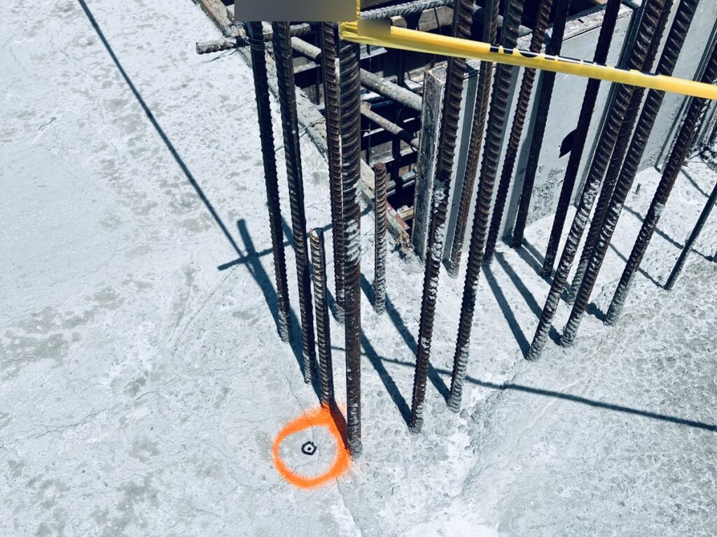 Marca de replanteo naranja en el suelo junto a una columna de varillas de acero en una obra