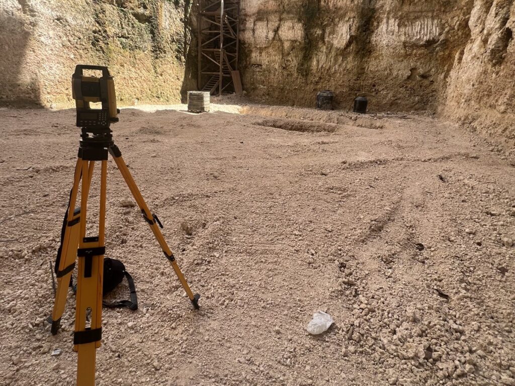 Estación total sobre trípode en un solar excavado, con edificios al fondo